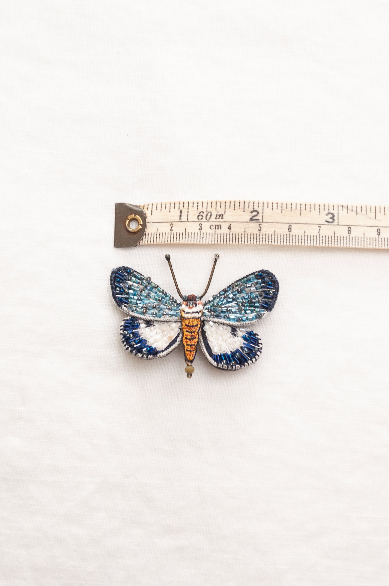 Trovelore Neochera moth hand beaded brooch, detailed butterfly-shaped brooch with iridescent blue and white wings, a metallic gold body, and delicate antennae.