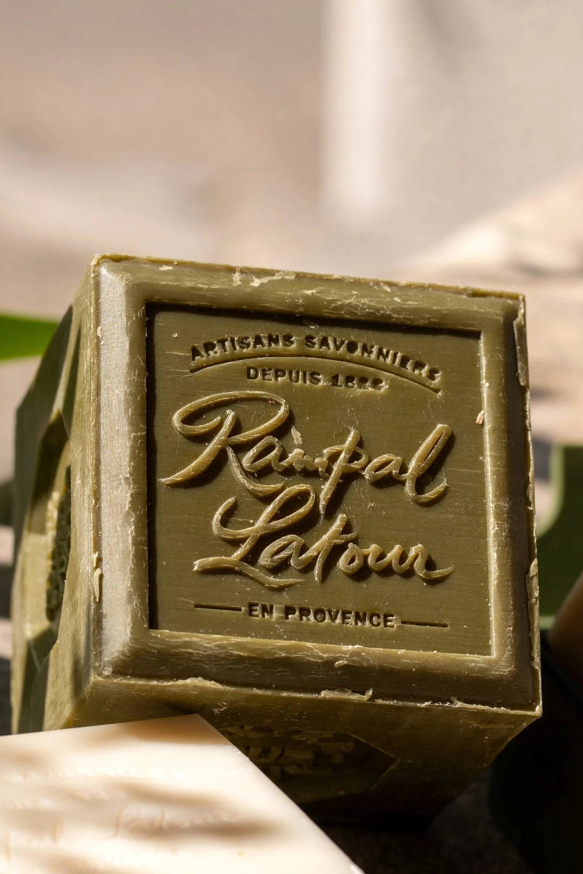 A rectangular bar of green soap with embossed lettering reading 'Rampal Lavoir' and 'Artisans Savonniers Depuis 1828 En Provence' on the front, photographed outdoors with sunlight and shadows.